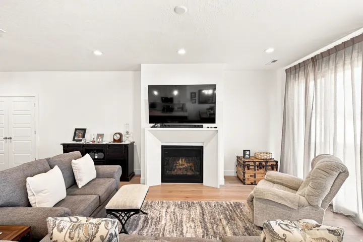Bright, open living room featuring a gas fireplace with built-in cabinetry, wall-mounted flat-screen television, recessed ceiling lighting, and expansive windows with floor-to-ceiling drapery