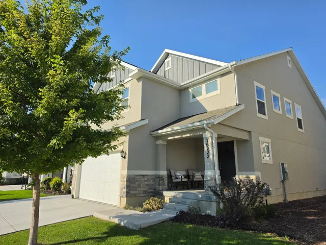 Contemporary two-story home featuring a taupe and stone exterior with mixed material detailing, covered front porch with stone accents, multi-pane windows, attached two-car garage, manicured landscaping with mature trees, and clean architectural lines wit