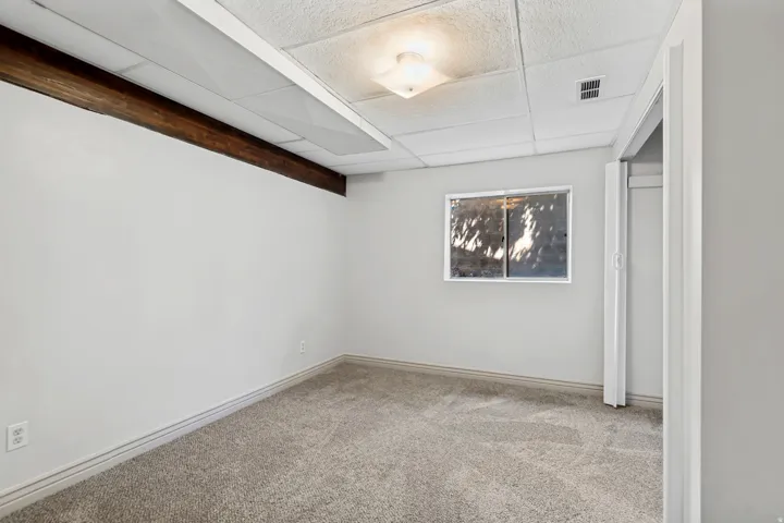Bright basement room with neutral carpeting, recessed ceiling lighting, casement window with natural light, exposed wood beam detail, and white trim throughout