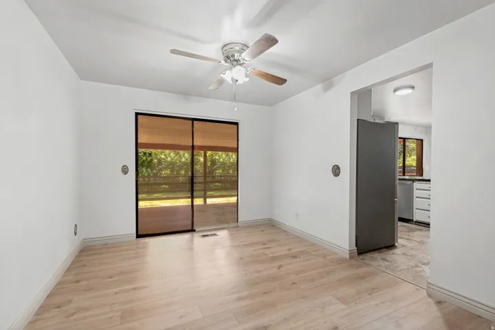 Bright, open living space featuring wood-finish flooring, sliding glass doors with bronze-tone frames, ceiling fan with integrated lighting, and direct access to the kitchen area