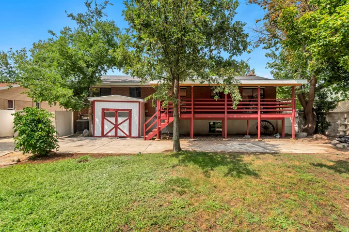 Expansive backyard featuring a red-painted deck, mature trees, a white and red storage shed, and a concrete patio
