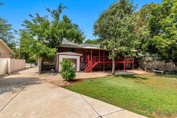 Expansive concrete driveway, detached storage shed with red trim, private deck with red railings, mature trees, and a grassy yard
