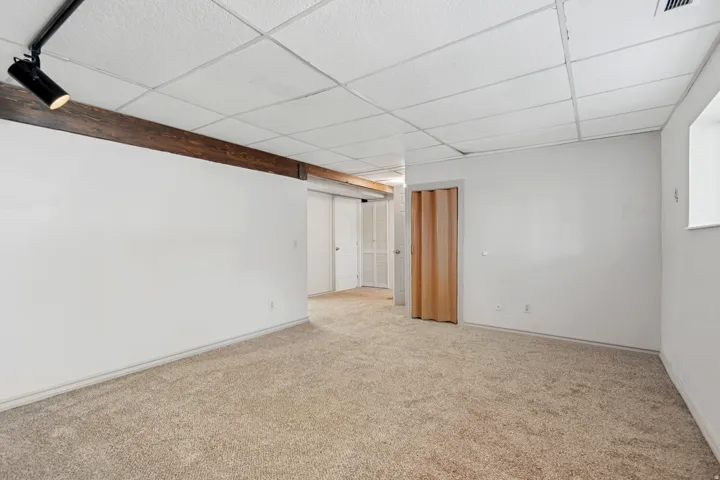 Carpeted room with white walls, wood beam accent, track lighting, and a drop ceiling