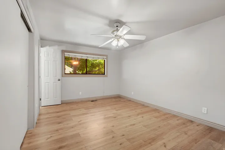 Wood-finish flooring, white ceiling fan, light gray wall paint, window with wood trim, and a white paneled door