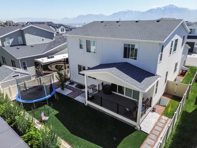 Covered deck with dark railings, paved patio with white stone, artificial turf lawn, privacy fencing, and exterior storage shed