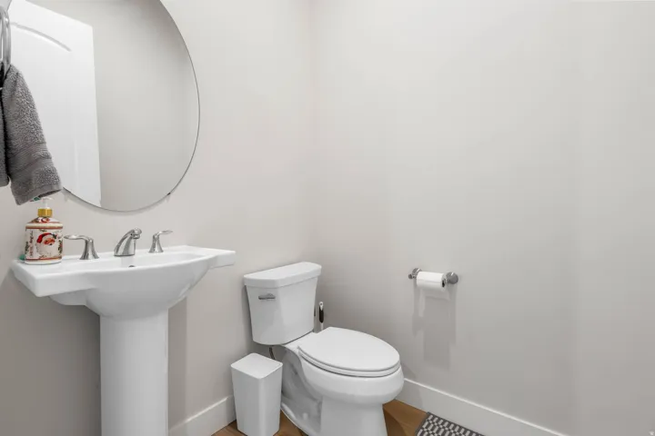 Pedestal sink with chrome faucet, circular wall mirror, white toilet, and wood-finish flooring