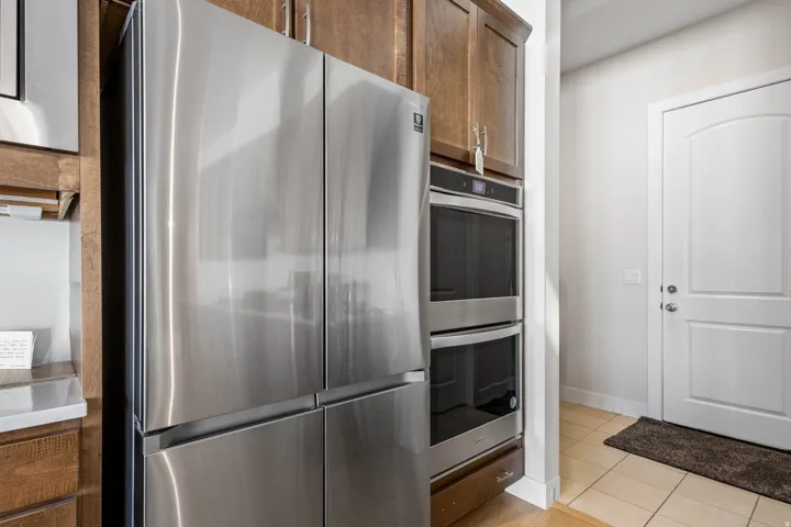 Stainless steel French door refrigerator, built-in double ovens, wood-finish cabinetry, light-toned tile flooring, and a white paneled door