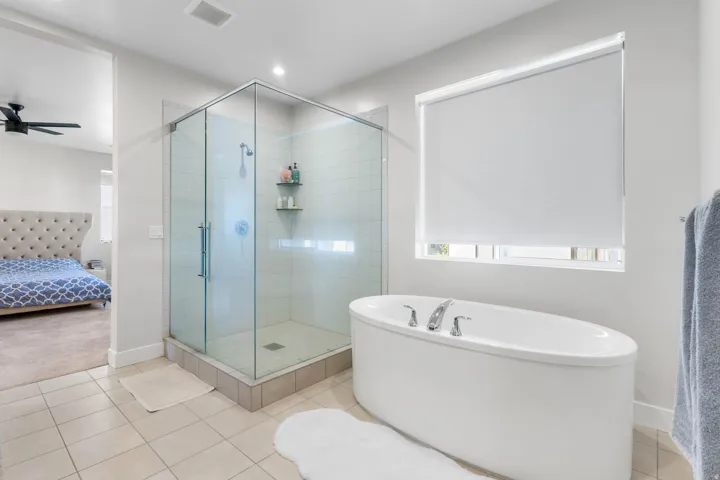 Glass-enclosed shower, freestanding soaking tub, tiled flooring, roller window shades, and recessed lighting