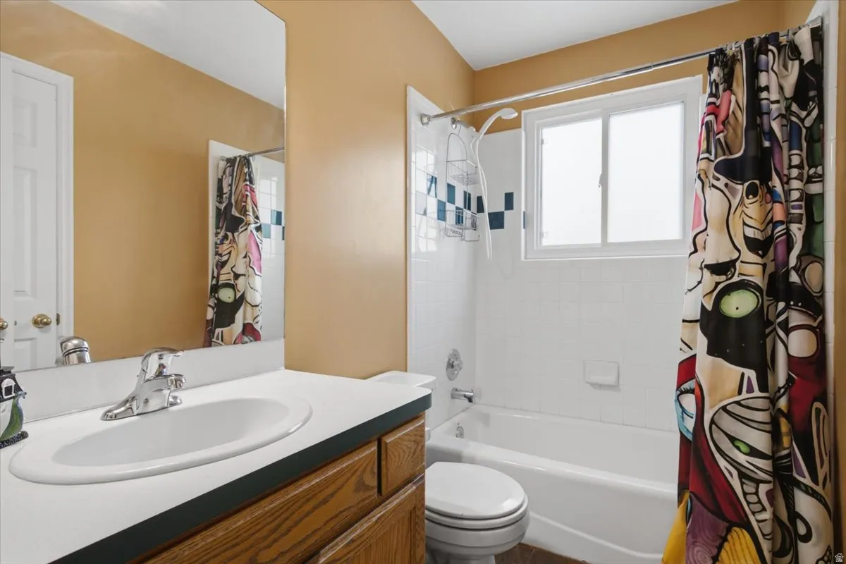 Bright bathroom featuring warm golden walls, a curved sink with wood-finish vanity cabinetry, white subway tile surround with glass enclosure shower/tub combination, and coordinating toilet