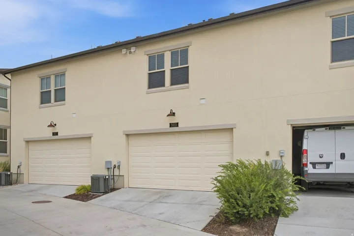 Modern multi-car garage featuring three white sectional doors with contemporary hardware, concrete driveway with clean finish, and mature landscaping with low-maintenance shrubs framing the entry