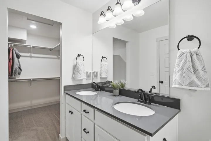 Dual vanity with integrated sinks and dark countertop, large mirrored surface, white cabinetry with dark hardware, and adjacent walk-in closet with built-in shelving