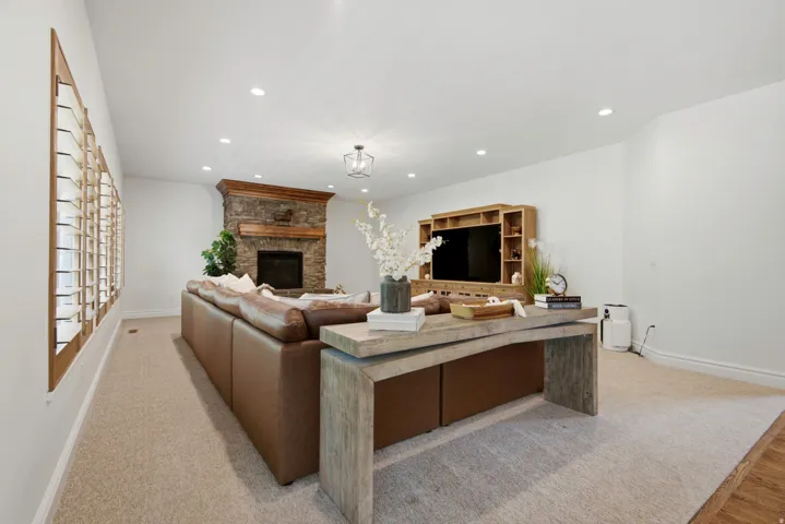 Recessed lighting, light-toned carpeting, wood-frame plantation shutters, stone fireplace surround with wood mantel, and a decorative ceiling fixture