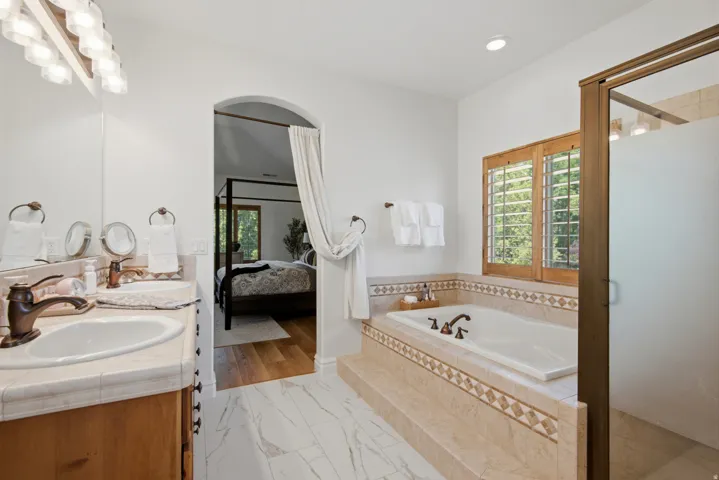 Dual vanity with integrated sinks, a built-in soaking tub, a separate shower enclosure, wood-finish window shutters, and marble-look flooring