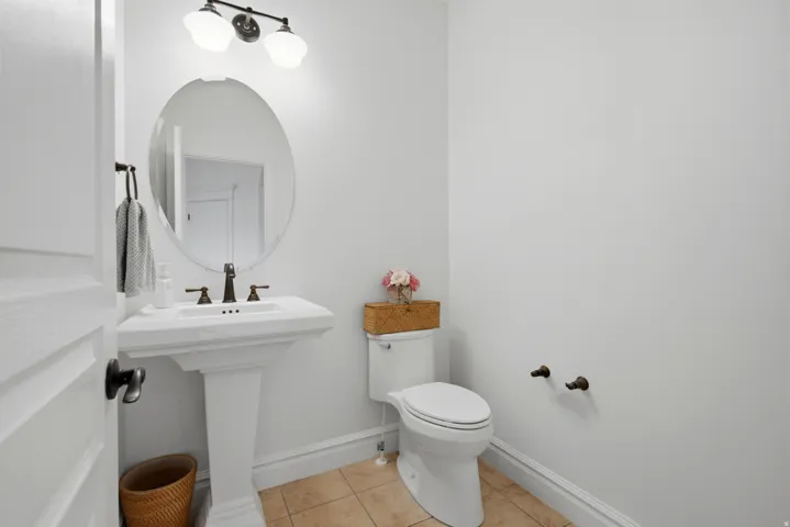 Pedestal sink with dark bronze faucet, oval mirror, wall-mounted light fixture, and tile flooring