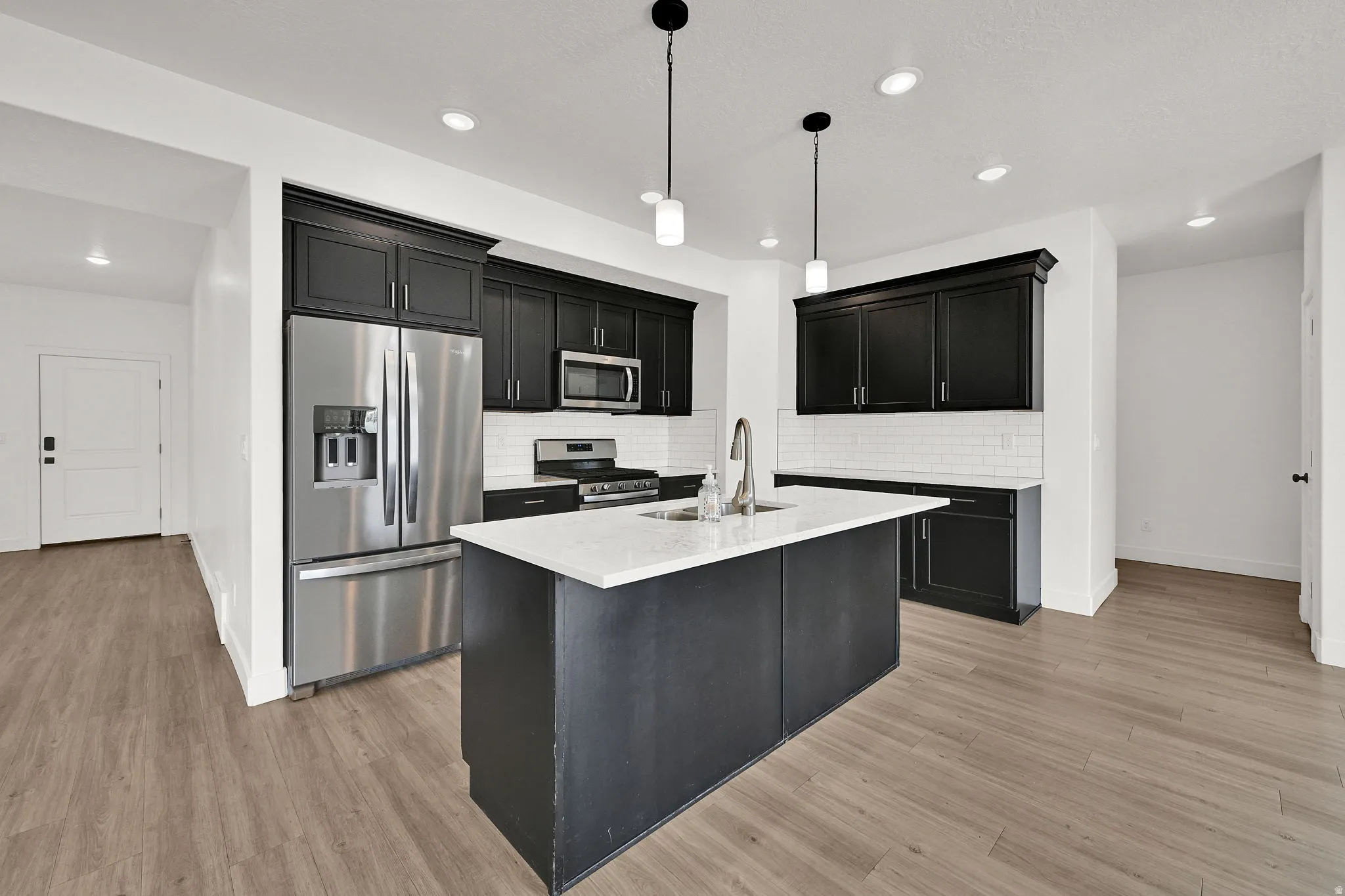 Wood-finish flooring throughout, stainless steel appliances, black cabinetry, white subway tile backsplash, and a central island with a solid surface countertop
