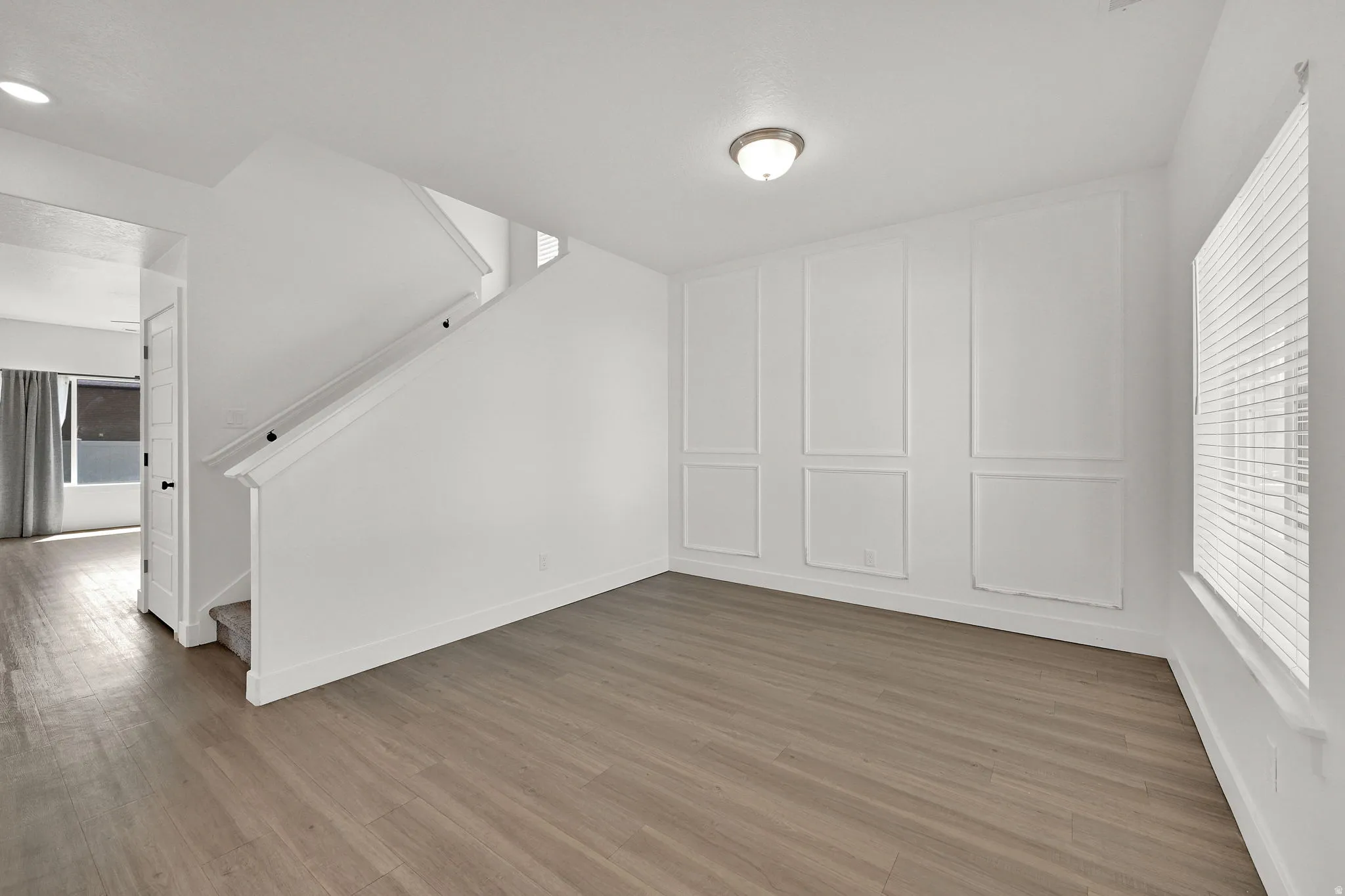 Wood-finish flooring, accent wall paneling, flush mount lighting, staircase with white railing, and window with blinds