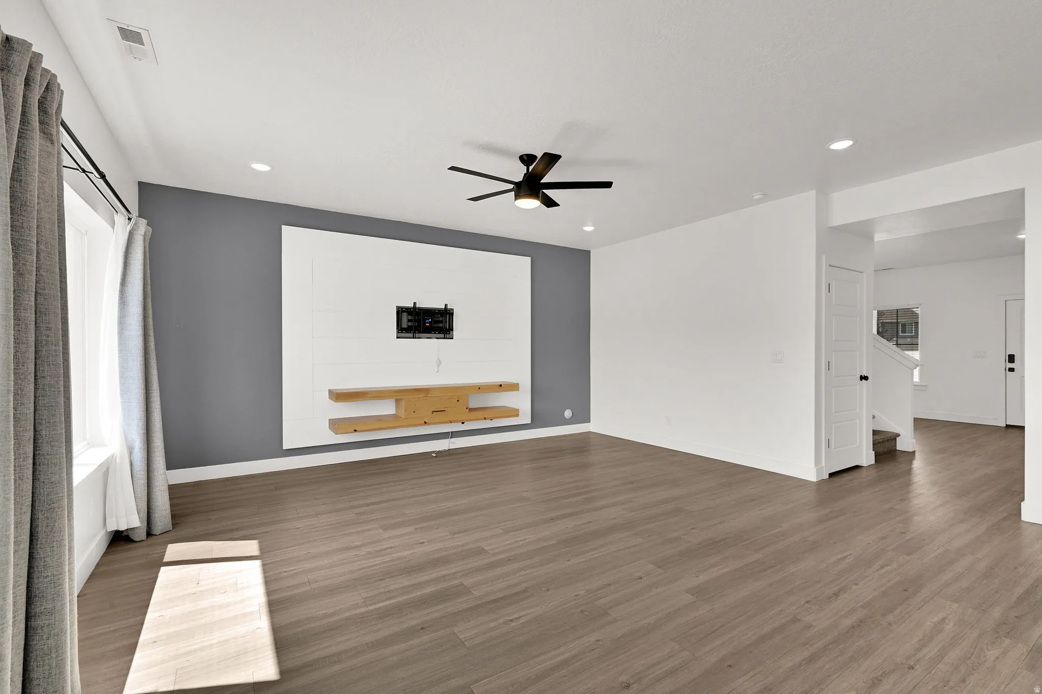 Wood-finish flooring, recessed lighting, ceiling fan, accent wall with built-in shelving, and window with drapes