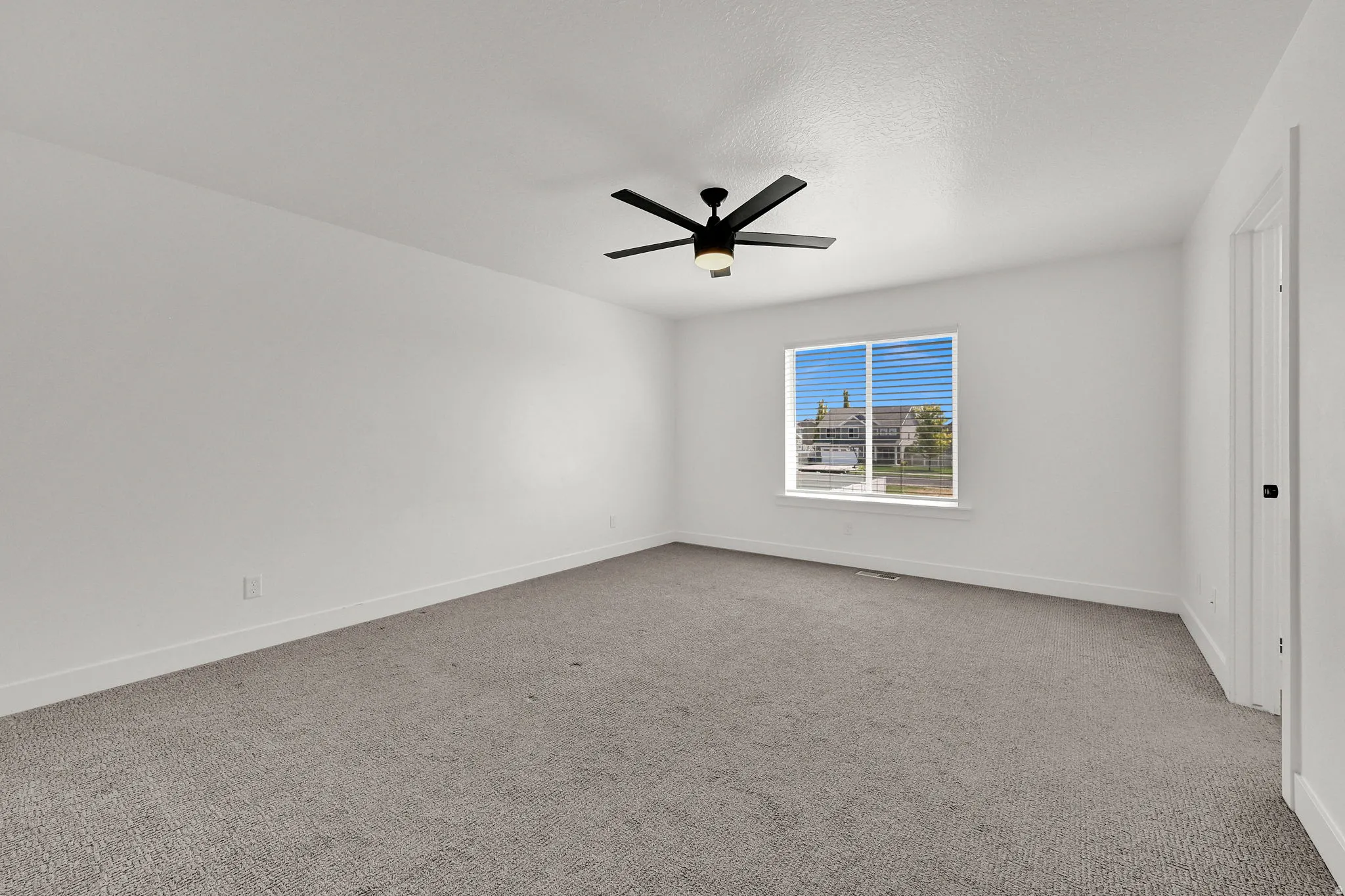 Expansive room with neutral carpeting, a large window, a modern ceiling fan, white walls, and a white door with black hardware