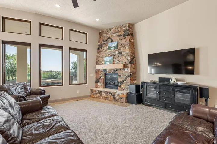 Stone-clad fireplace with wood mantel, large windows with upper transom lights, recessed lighting, and a ceiling fan