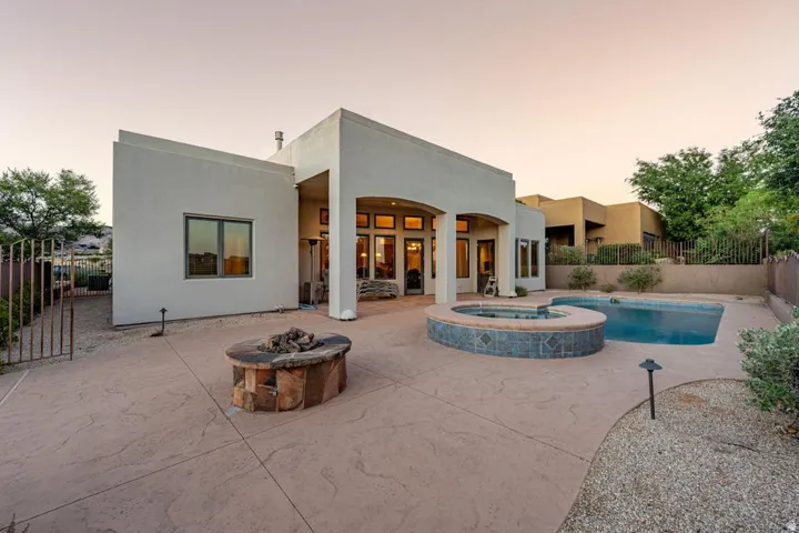 Modern architectural home with expansive concrete patio featuring a resort-style saltwater pool with integrated spa, fire pit with seating area, mature desert landscaping with native vegetation, and covered outdoor living space with arched entryway and la