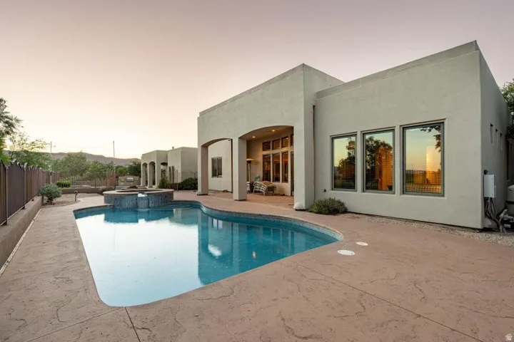 Curved swimming pool with integrated spa and waterfall feature, stamped concrete patio, stucco exterior, and multi-pane windows