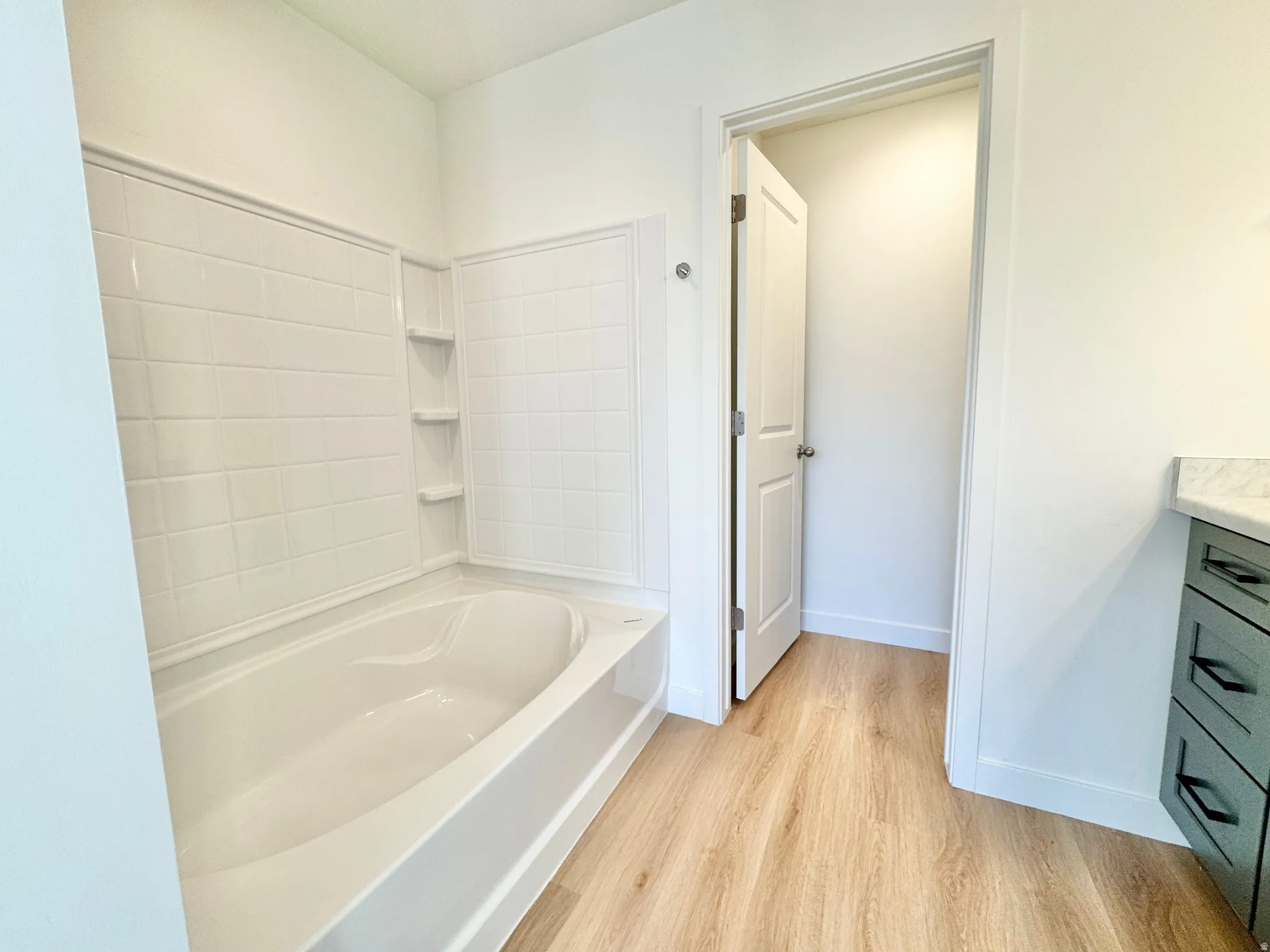Bathtub with white subway tile surround, built-in shelving, wood-finish flooring, and a vanity with dark hardware and a light countertop