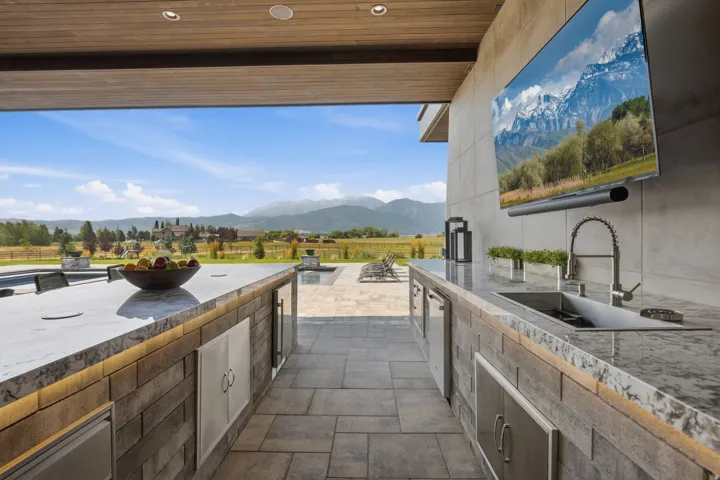 Expansive outdoor kitchen featuring stone counters, integrated sink with gooseneck faucet, built-in appliances, and a wall-mounted display