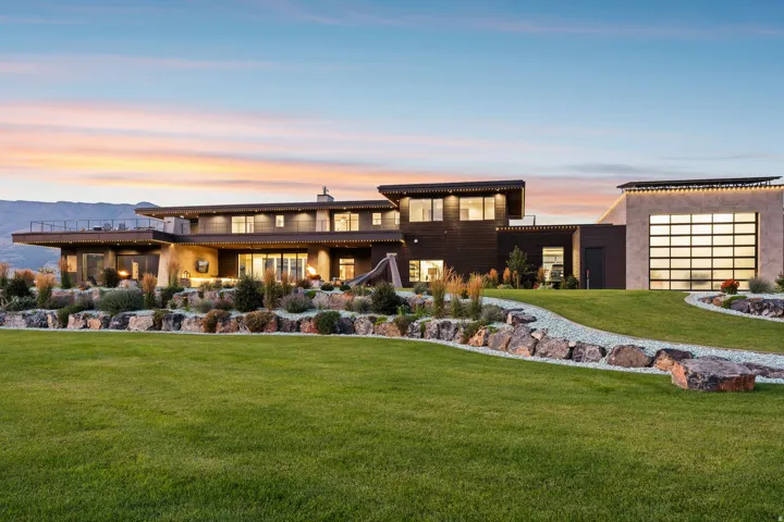 Modern exterior architecture with multiple levels, expansive lawn, terraced landscaping with rock accents, a large glass-paneled garage door, and a cantilevered deck