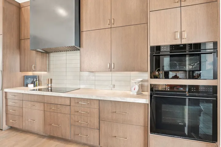 Integrated double ovens, built-in cooktop, stainless steel range hood, light wood-finish cabinetry, and a subway tile backsplash