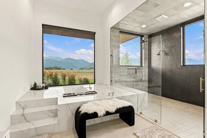 White soaking tub integrated into a marble-look surround with steps, large picture window with scenic backdrop, glass-enclosed shower with dark textured wall tile, recessed shelving, and a ceiling-mounted shower head