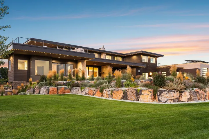 Expansive green lawn with a rock retaining wall, mature landscaping, and an exterior featuring large windows and multiple outdoor living areas