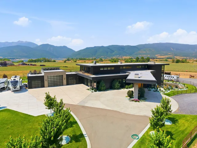 Contemporary exterior with dark siding, large windows, an expansive paved driveway, multiple garage doors, and surrounding mountain scenery
