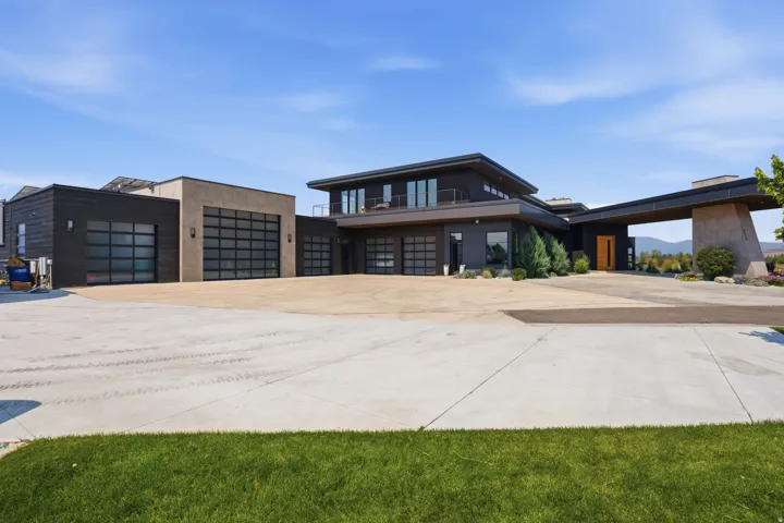 Modern architectural design featuring expansive concrete driveways, multiple garage bays with frosted glass doors, a prominent porte-cochère entrance, and a second-story balcony