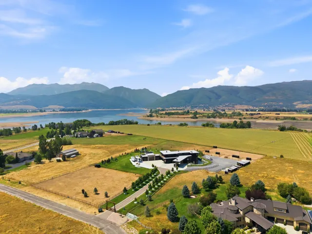 Expansive property featuring a contemporary residence, additional structures, natural landscape, and a paved driveway, all set against a backdrop of mountains and a large body of water