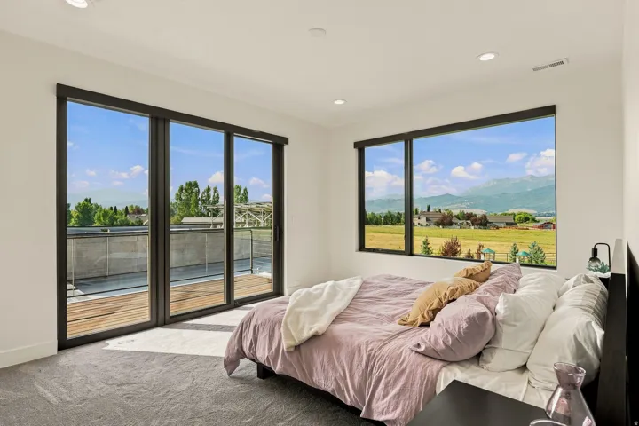 Large windows, recessed ceiling lights, dark-framed sliding glass doors, exterior balcony with wood-finish decking, and grey carpeting