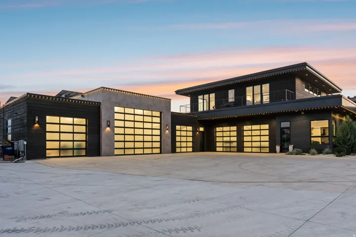 Modern exterior featuring dark wood-finish siding, multiple full-light garage doors, extensive concrete hardscaping, and upper-level balconies with metal railings