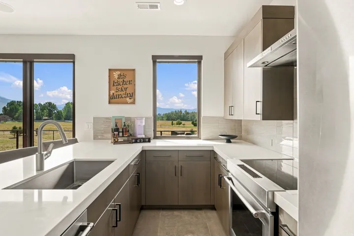 Modern kitchen featuring light wood-finish cabinetry, white countertops, stainless steel appliances, a tile backsplash, and two large windows