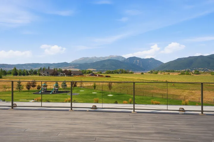 Expansive outdoor deck with glass panel railing overlooking a landscaped green space, distant mountains, and agricultural fields