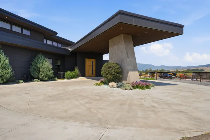 Modern architecture featuring a dark exterior, a prominent porte-cochère, a wood-finish entry door, and a concrete driveway