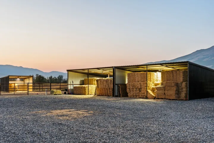 Gravel drive surface, mountain backdrop, metal-sided storage structures with open fronts, interior lighting, and metal fencing