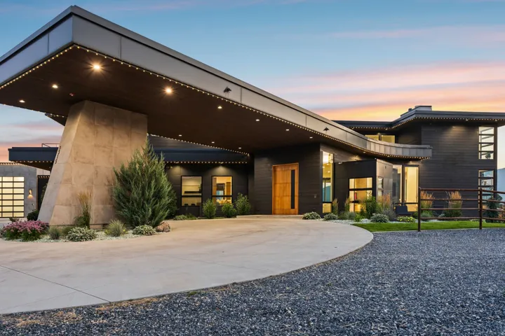 Modern architectural exterior featuring dark siding, a large covered entry with recessed lighting, an expansive concrete driveway, and a visible garage door