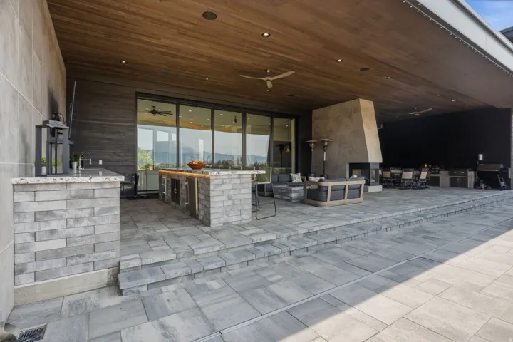 Expansive covered patio featuring a wood-finish ceiling, stone outdoor kitchen with built-in appliances, recessed lighting, and a prominent fireplace with a substantial hearth