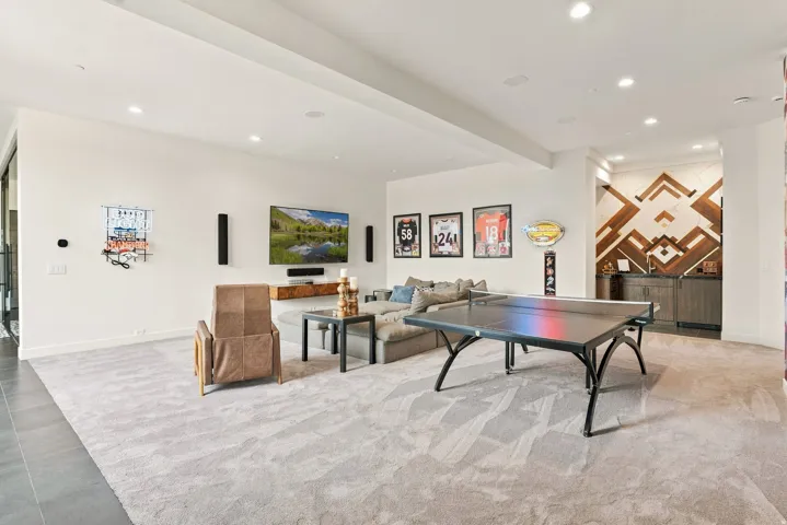 Recessed lighting illuminates the spacious room featuring light-toned carpeting and a built-in wet bar with a decorative wood-panel accent wall