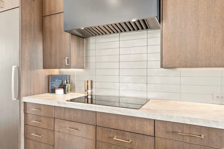 Wood-finish cabinetry with gold-tone hardware, a light-toned countertop, and a sleek cooktop with a coordinating range hood