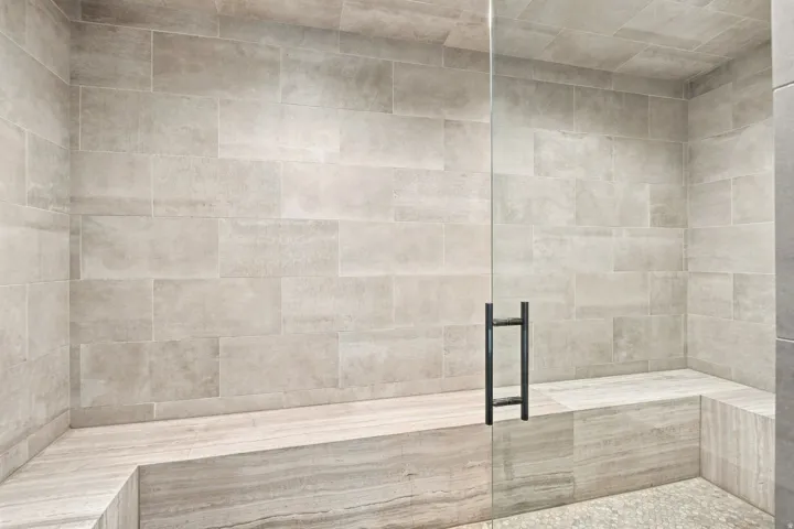 Tiled shower enclosure featuring a glass panel door with a matte black handle, a built-in bench, and a river rock-style floor