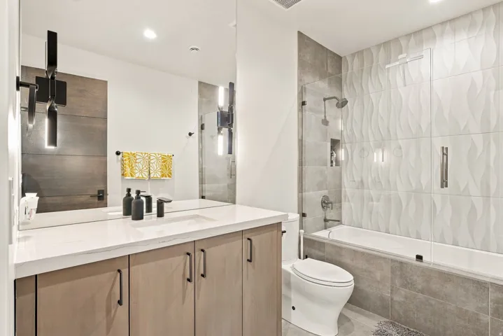 Contemporary bathroom featuring a light wood-finish vanity with a white countertop, a built-in bathtub with glass enclosure, a shower with a tiled surround, and textured wall tiles