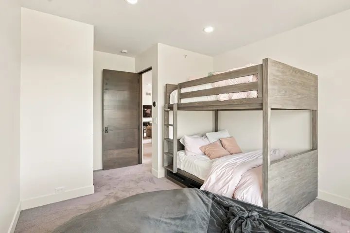Modern bedroom suite featuring a wood-finish loft bed with horizontal slat railings, recessed ceiling lighting, neutral wall finishes, and direct access to an ensuite bathroom with tile shower surround and brushed metal fixtures