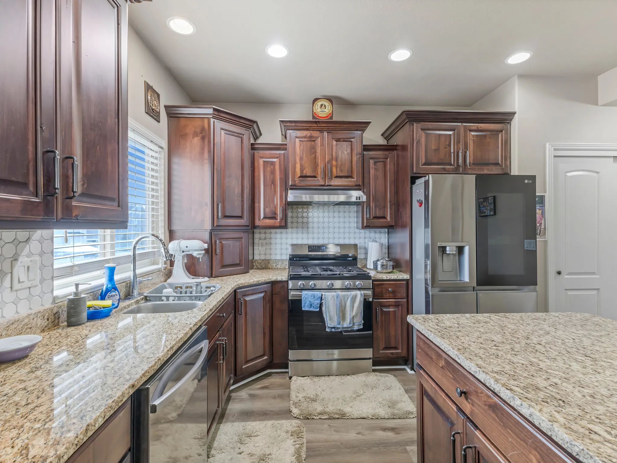 Kitchen featuring rich wood cabinetry, light granite countertops, stainless steel appliances, a tile backsplash, and recessed lighting