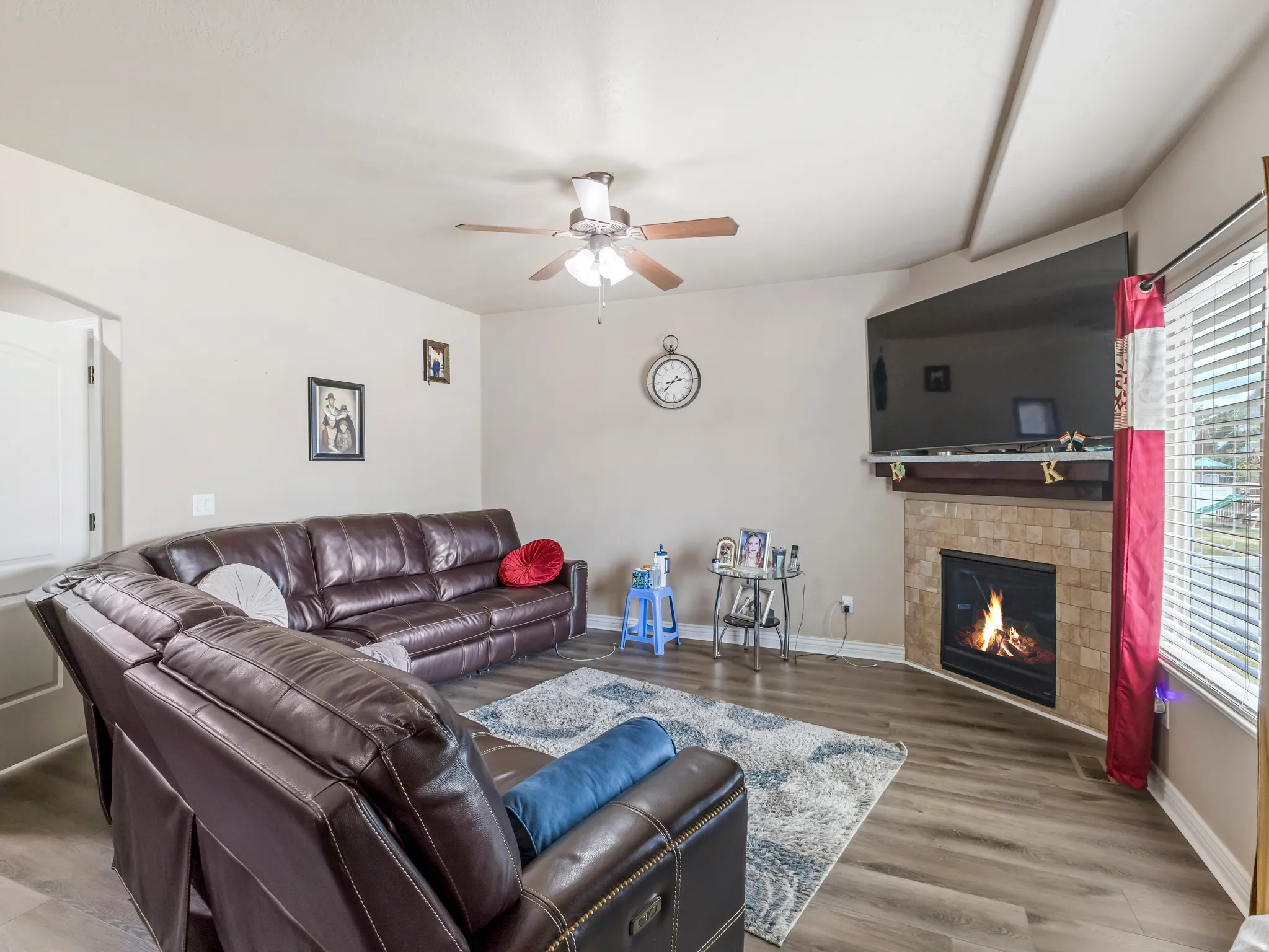 Living area featuring wood-finish flooring, a ceiling fan, a tiled fireplace with a mantel, and window blinds