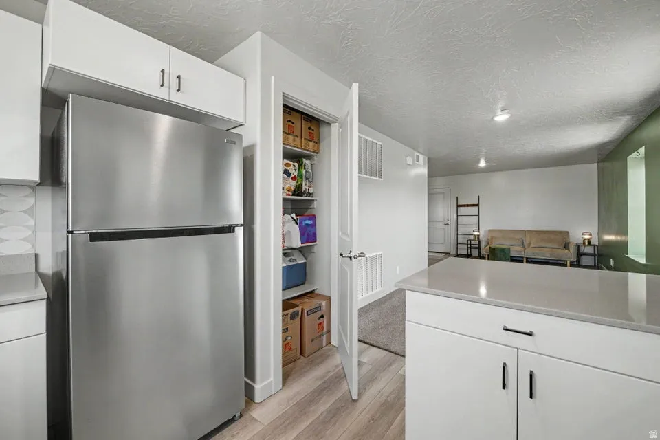 Stainless steel refrigerator, white cabinetry, light wood-finish flooring, light-colored countertops, and pantry storage